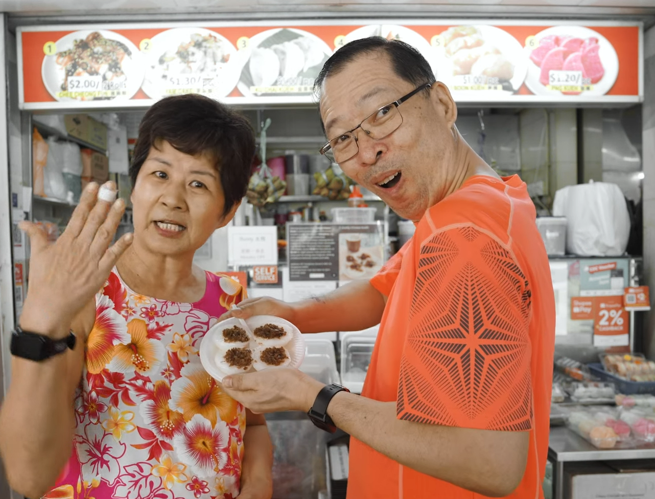 Two people posing at a food stall while holding a plate of chwee kueh, capturing a lively and cheerful moment in a hawker centre setting.